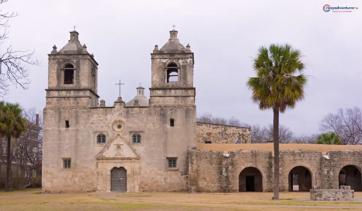 A historic Spanish mission with two bell towers made of stone stands before a cloudy sky. A single palm tree is on the right, and a row of stone arches leads to a courtyard. The scene is calm and deserted.
