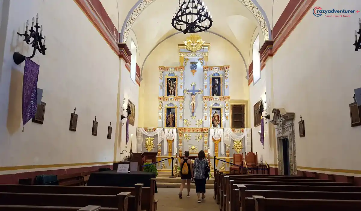 Two people walk towards the ornate altar in a historic church with high ceilings. The altar features religious statues, a crucifix, and decorative elements. Wooden pews line the aisle, and chandeliers hang from the ceiling.
