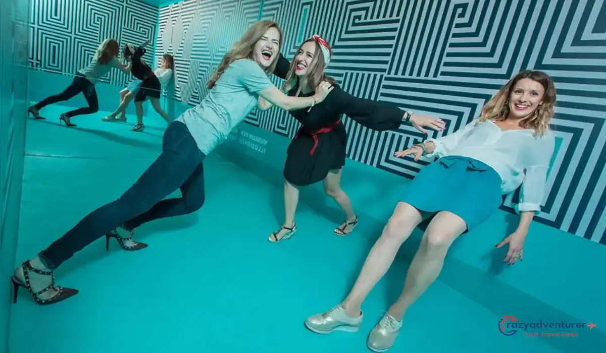 The 10 Best Interactive Museums in NYC You Must Visit  9 Three women pose playfully in a room with optical illusions, giving the appearance of leaning at impossible angles. The room has a blue floor and patterned walls, creating a disorienting effect.