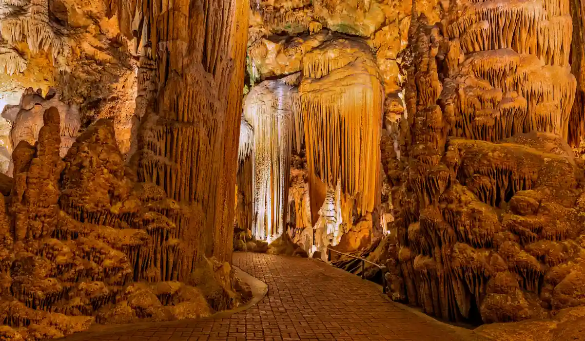A breathtaking cave with large stalactites and stalagmites made of orange and brown rock formations. A paved pathway winds through the illuminated cavern, showcasing the intricate natural structures.

