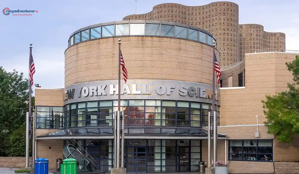 The 10 Best Interactive Museums in NYC You Must Visit  6 Exterior view of the New York Hall of Science. The building features a cylindrical structure with large glass windows and three American flags at the entrance. The facade is made of light-colored bricks and there is greenery nearby.