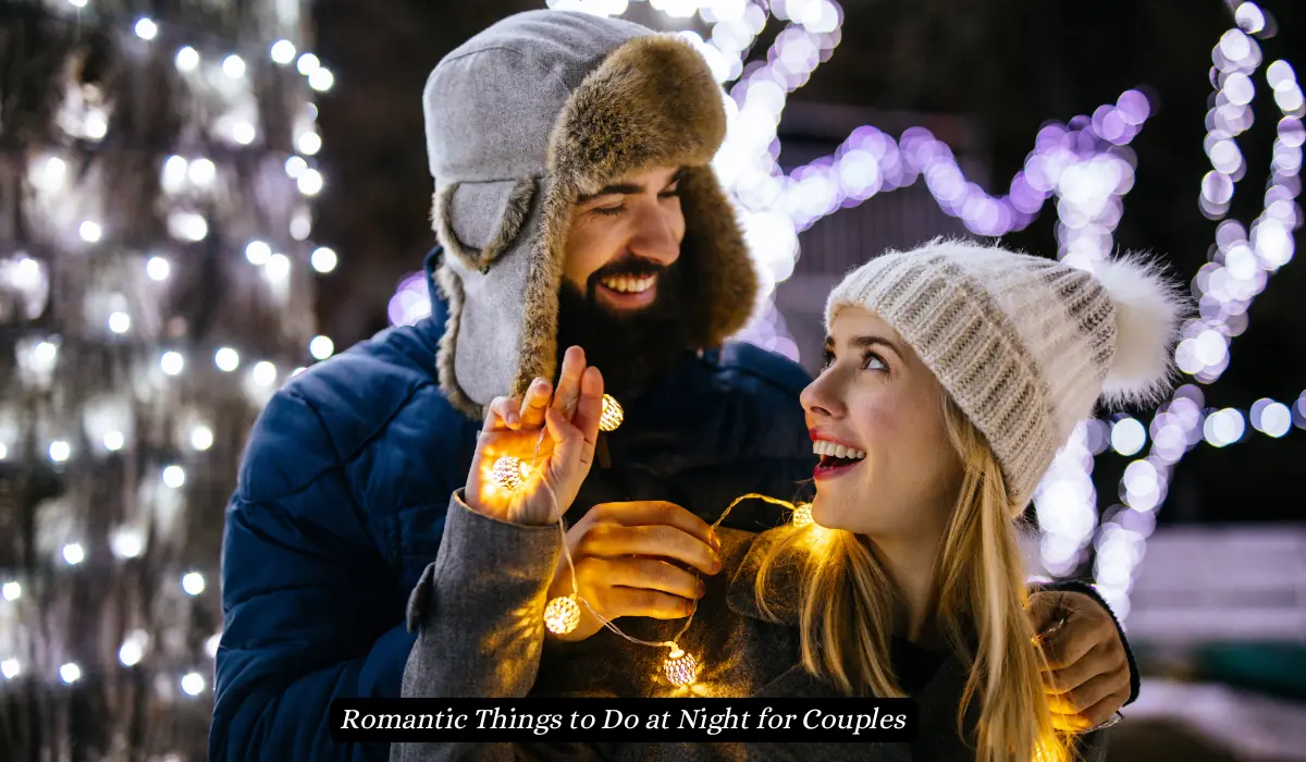 A couple in warm clothing is playfully wrapped in string lights, smiling at each other outdoors at night. The background features softly glowing white lights, creating a festive and romantic atmosphere. "Romantic Things to Do at Night for Couples"