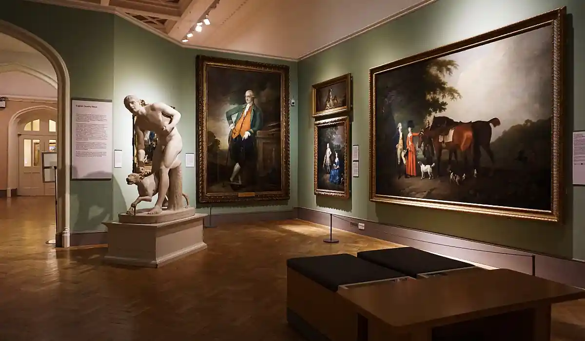 A museum gallery featuring a white statue of a standing male figure with a dog at his feet, surrounded by several large framed paintings on olive green walls. A wooden bench is positioned in the center of the room.
