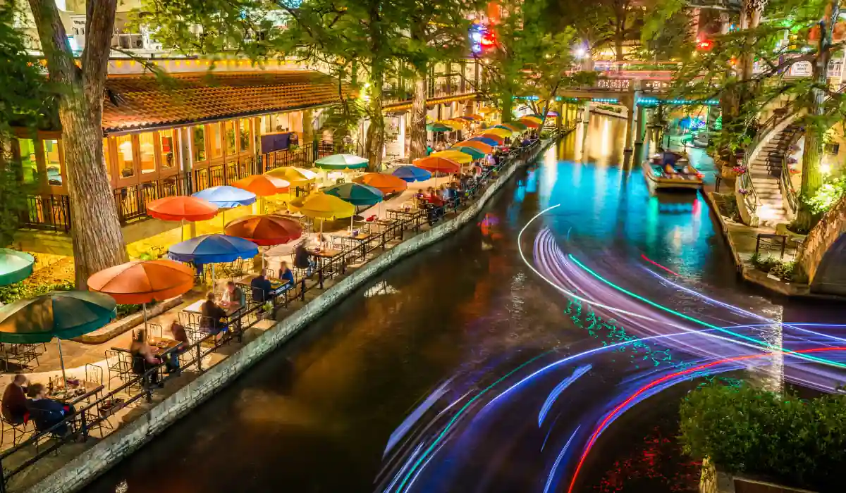 A vibrant night scene of a riverwalk with colorful umbrellas lining the waters edge, reflecting bright lights. People sit along the waterfront, and streaks of lights from passing boats create dynamic patterns on the water.
