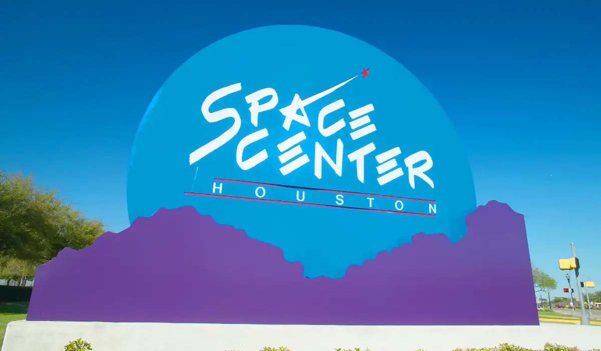 15+ Fun and Romantic Things To Do In Houston for Couples 3 A large blue and purple sign for Space Center Houston against a clear blue sky. The words Space Center Houston are displayed in bold white letters, with a red arrow motif. Trees and a streetlamp are visible in the background.