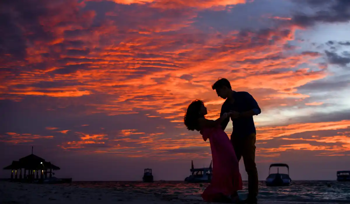 Top 15 Best Things to do in Gatlinburg TN for Couples 11 A couple in silhouette poses romantically on a beach at sunset, with vibrant orange and purple clouds filling the sky. Boats are visible in the water, and a small building is seen on the left.