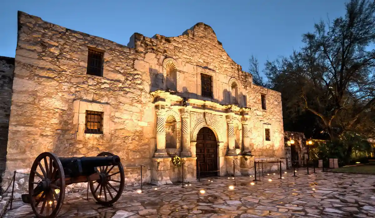 A historic stone building with arched wooden doors is illuminated at dusk. A cannon is positioned in the foreground on a cobblestone plaza. Trees are visible in the background under a clear blue sky.
