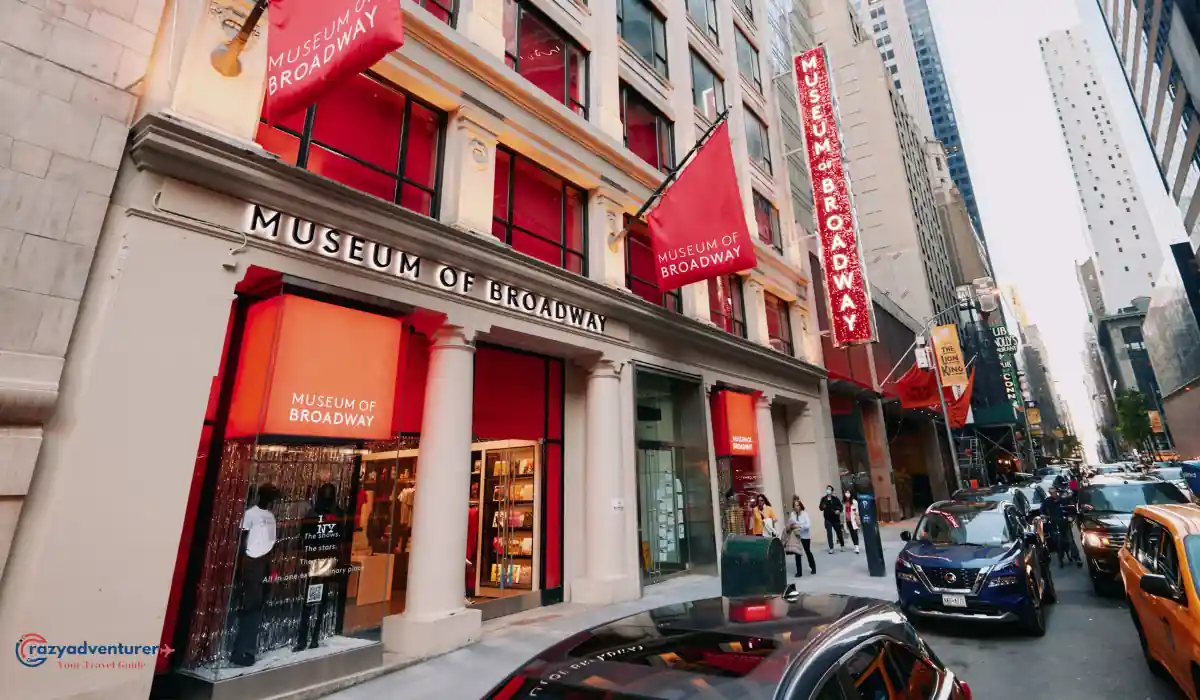 The 10 Best Interactive Museums in NYC You Must Visit  2 Street view of the Museum of Broadway with large red banners and multiple entrances. People are walking on the sidewalk, and cars are parked along the street. The building features classic architecture with columns and red accents.