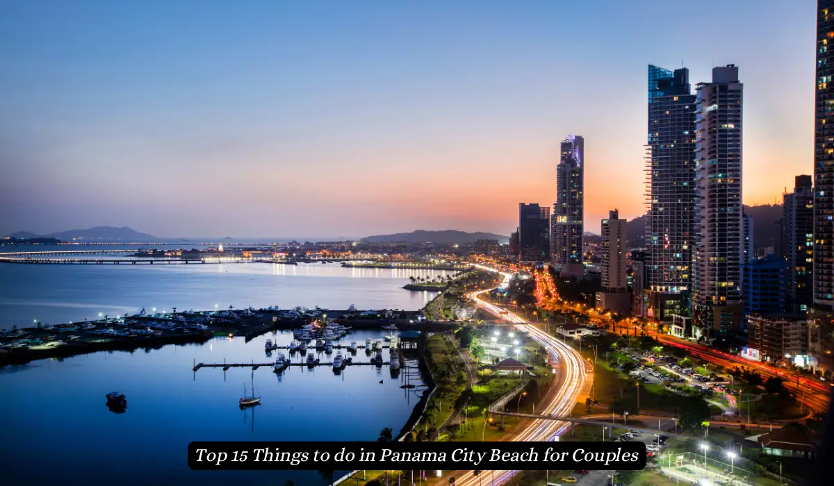 Aerial view of a coastal cityscape at dusk, with illuminated skyscrapers lining the waterfront. A busy highway with light trails leads through the city, and boats are docked in a marina. Lush greenery and a distant mountain complete the scene. "Things to do in Panama City Beach for Couples"