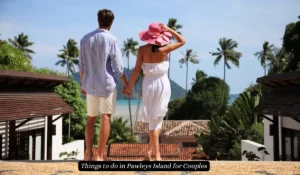 A couple holding hands, facing towards a tropical view with palm trees and the ocean. The woman wears a pink sunhat and white dress, while the man is in a light shirt and shorts. The text reads, Things to do in Pawleys Island for Couples.