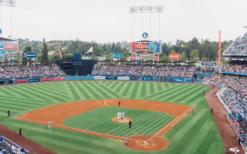 Top 18 Best Things to Do in San Jose for Couples 3 A baseball game in progress at a large stadium. The field shows neatly mowed grass with a dirt infield. Players are gathered on the pitchers mound, surrounded by an audience in the stands under a cloudy sky. Advertising banners are visible.