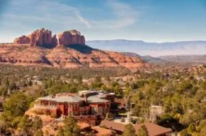 A large, elegant house surrounded by trees sits in the foreground, with dramatic red rock formations and mesas rising in the background under a clear blue sky.