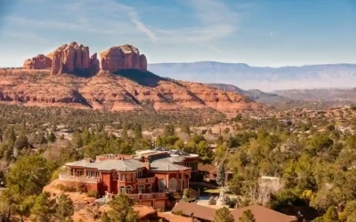 A large, elegant house surrounded by trees sits in the foreground, with dramatic red rock formations and mesas rising in the background under a clear blue sky.