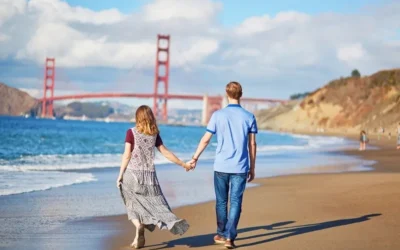 A couple holds hands while walking along a sandy beach with the Golden Gate Bridge in the background under a partly cloudy sky.