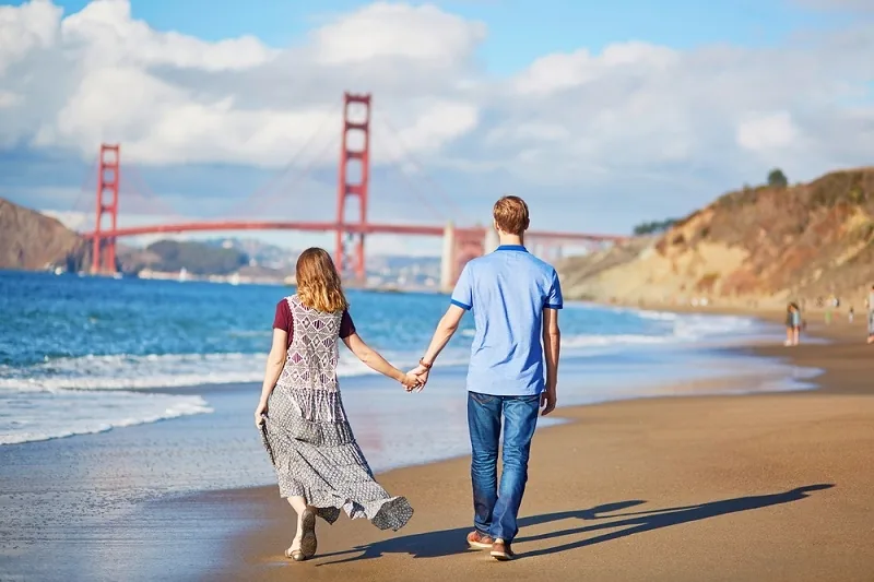 A couple holds hands while walking along a sandy beach with the Golden Gate Bridge in the background under a partly cloudy sky.