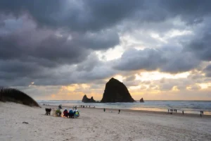 People sit around a beach fire on sandy shore as others walk nearby, with Haystack Rock rising from the ocean under a dramatic, cloudy sunset sky at Cannon Beach, Oregon.