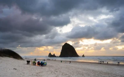 People sit around a beach fire on sandy shore as others walk nearby, with Haystack Rock rising from the ocean under a dramatic, cloudy sunset sky at Cannon Beach, Oregon.
