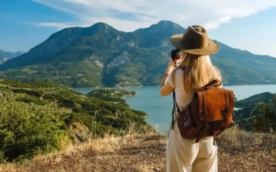 The Rise of Solo Travelers CreativeCultureTribe: 8 Secrets A woman with a brown backpack and wide-brim hat stands on a grassy hill, taking a photo of a scenic landscape with mountains, a lake, and greenery under a partly cloudy sky. The Rise of Solo Travelers CreativeCultureTribe 8 Secrets
