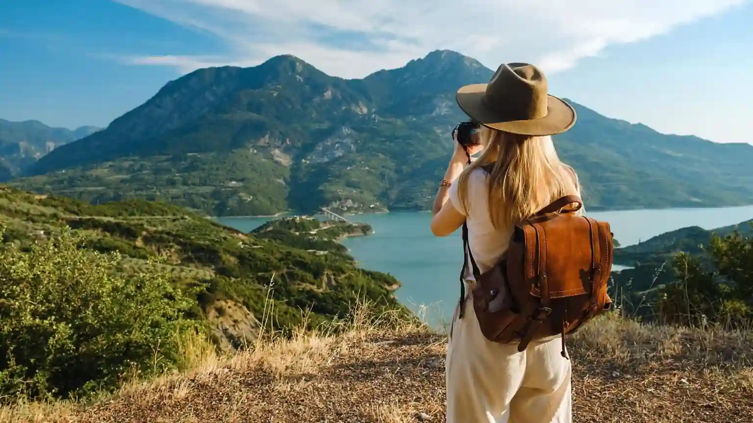 A woman with a brown backpack and wide-brim hat stands on a grassy hill, taking a photo of a scenic landscape with mountains, a lake, and greenery under a partly cloudy sky. The Rise of Solo Travelers CreativeCultureTribe 8 Secrets