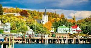 A scenic view of a lakeside village with colorful autumn trees, houses with green and white roofs, a church with a tall steeple, and a wooden pier in the foreground under a partly cloudy sky.
