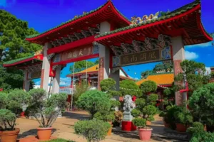 A traditional Chinese temple entrance with ornate red and green gates, Chinese characters, and stone lion statues, surrounded by lush green plants and trees under a bright blue sky. Things to Do in Houston Chinatown 9 Must-Visit Gems