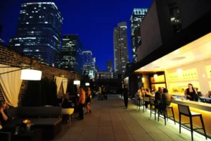 People sit and stand on a rooftop bar at night, surrounded by tall city buildings with lights, under a deep blue sky.