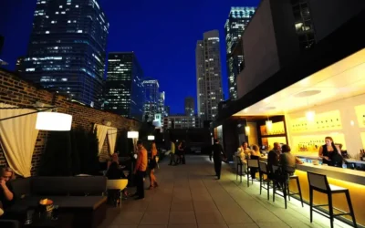People sit and stand on a rooftop bar at night, surrounded by tall city buildings with lights, under a deep blue sky.
