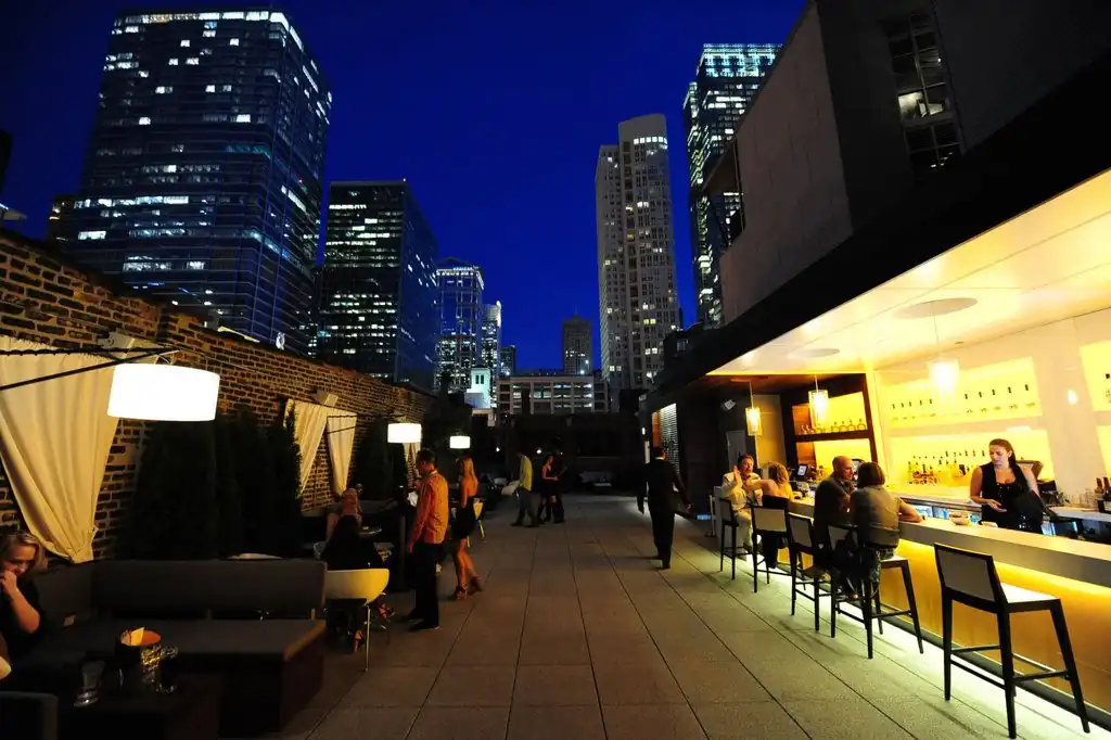 Top 15 Best Rooftop Bars in Chicago for Date Night Spots (2025) 56 People sit and stand on a rooftop bar at night, surrounded by tall city buildings with lights, under a deep blue sky.