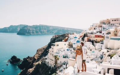 A couple stands on a white rooftop overlooking the sea and the iconic white-washed buildings with blue domes of Santorini, Greece, with cliffs and blue water visible in the background on a sunny day.