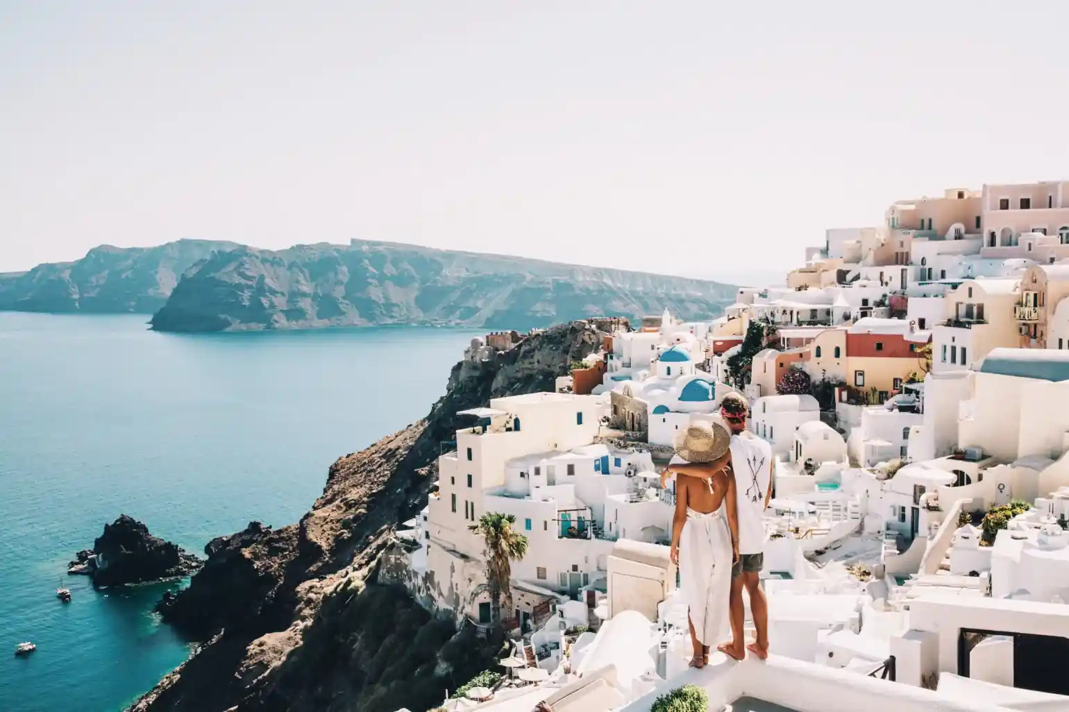 Top 20 Best & Dreamy Spots in Cincinnati for Couples 57 A couple stands on a white rooftop overlooking the sea and the iconic white-washed buildings with blue domes of Santorini, Greece, with cliffs and blue water visible in the background on a sunny day.