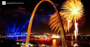 The image shows the St. Louis Gateway Arch at night with bright fireworks lighting up the sky and colorful spotlights shining over the city and river below.