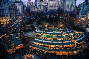Aerial view of an outdoor holiday market at dusk with green-roofed stalls, warm lights, crowds of people, and city skyscrapers in the background.