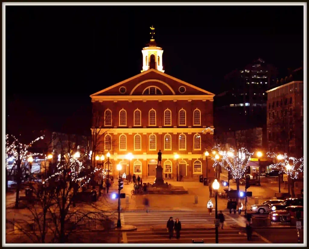 A warmly lit historic brick building with a clock tower, illuminated at night, stands in a plaza surrounded by trees adorned with string lights and people walking nearby.