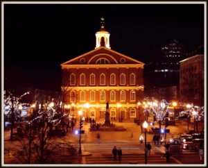 A warmly lit historic brick building with a clock tower, illuminated at night, stands in a plaza surrounded by trees adorned with string lights and people walking nearby.