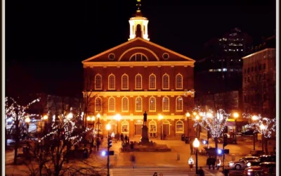 A warmly lit historic brick building with a clock tower, illuminated at night, stands in a plaza surrounded by trees adorned with string lights and people walking nearby.